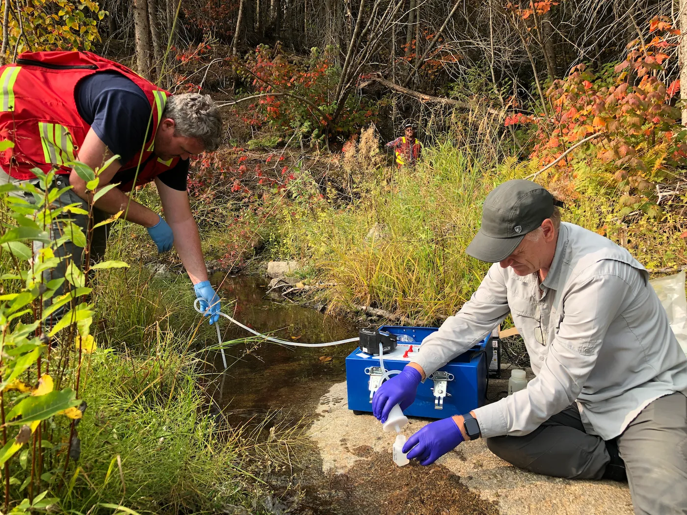 Two geochemists collecting and filtering a stream water sample in an autumn forest, using a portable field filtration pump